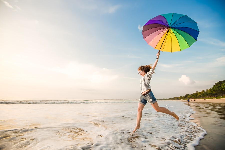 Cheerful young girl with rainbow umbrella having fun on the beach before sunset. Travel, holidays, v